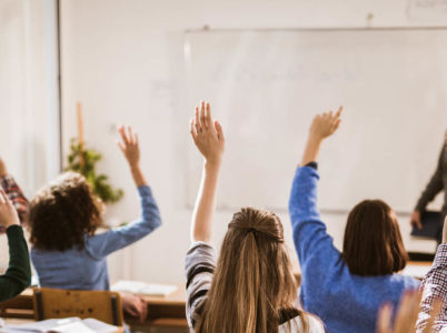 Photo of a class with teacher at the front, pointing at a student among many with their hands up